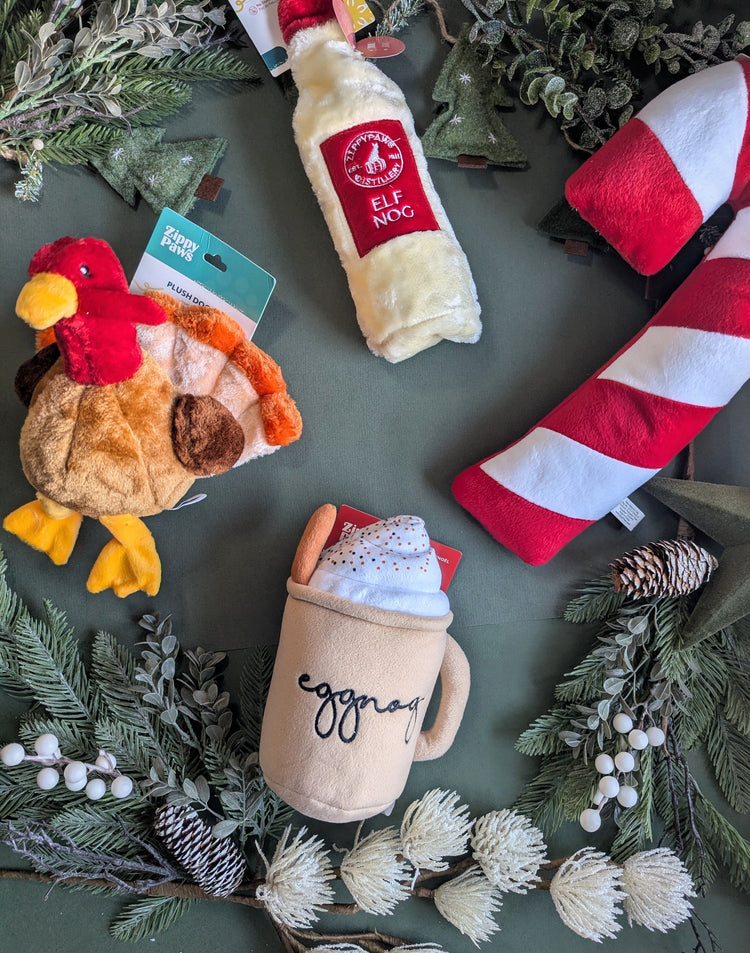 A variety of Christmas toys are laid out in a flat lay style, surrounded by Christmas foliage and pinecones, photographed on a dark green background