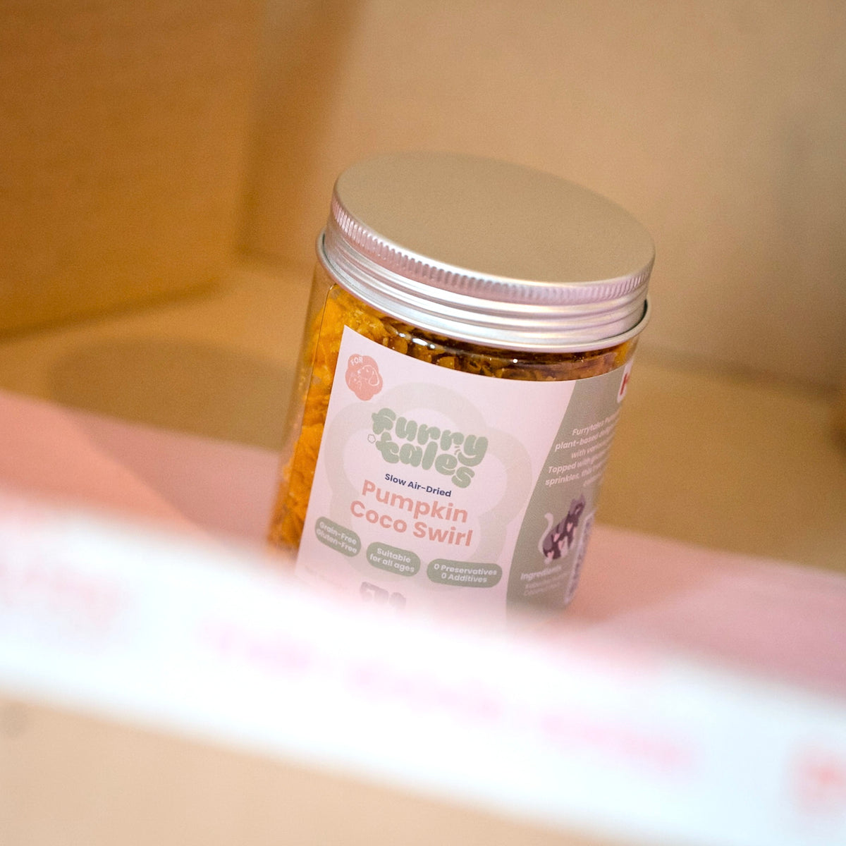 A clear, plastic jar of 'Furry Tales' Pumpkin Coco Swirl pet treats, set against a soft beige and pink background.