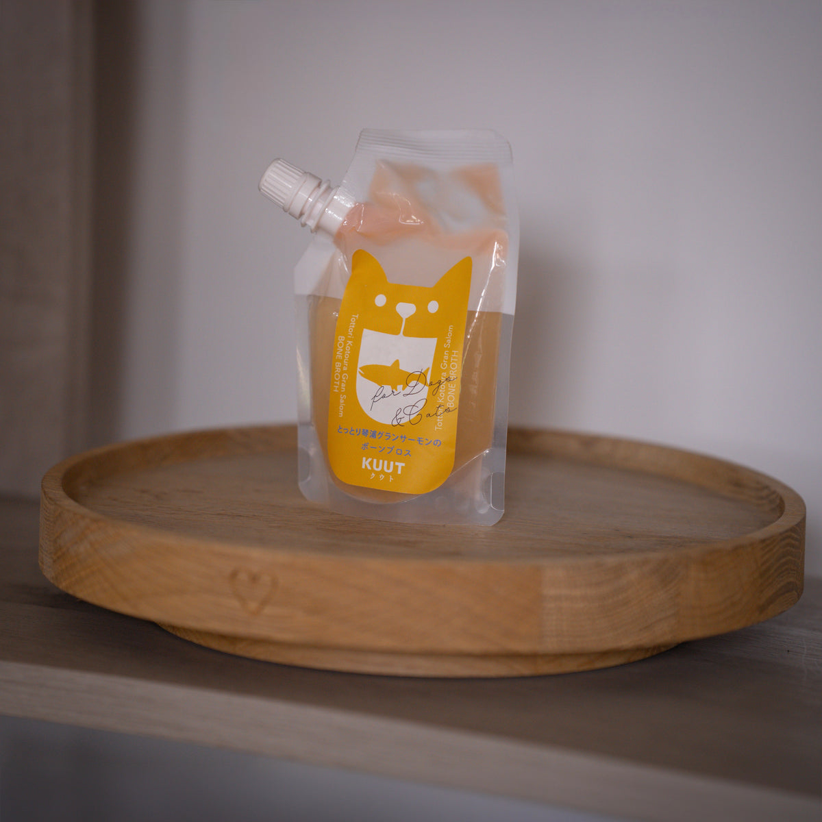 A clear plastic pouch of KUUT bone broth for dogs with a bright yellow cat-shaped label, resting on a round wooden tray against a neutral grey background.