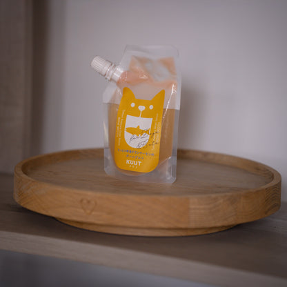 A clear plastic pouch of KUUT bone broth for dogs with a bright yellow cat-shaped label, resting on a round wooden tray against a neutral grey background.