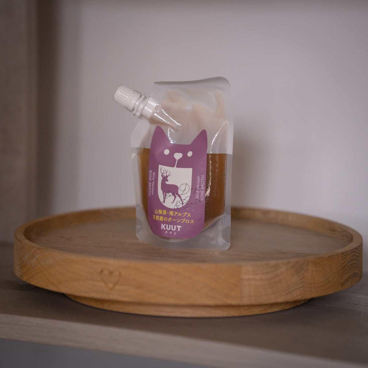 A clear stand-up pouch with a white cap and a purple cat-shaped label reading KUUT, containing bone broth from Japan, displayed on a round wooden tray against a soft neutral background.