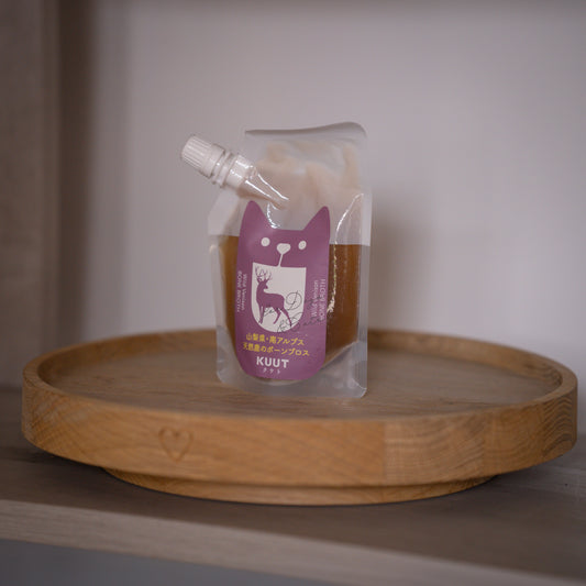 A clear stand-up pouch with a white cap and a purple cat-shaped label reading KUUT, containing bone broth from Japan, displayed on a round wooden tray against a soft neutral background.