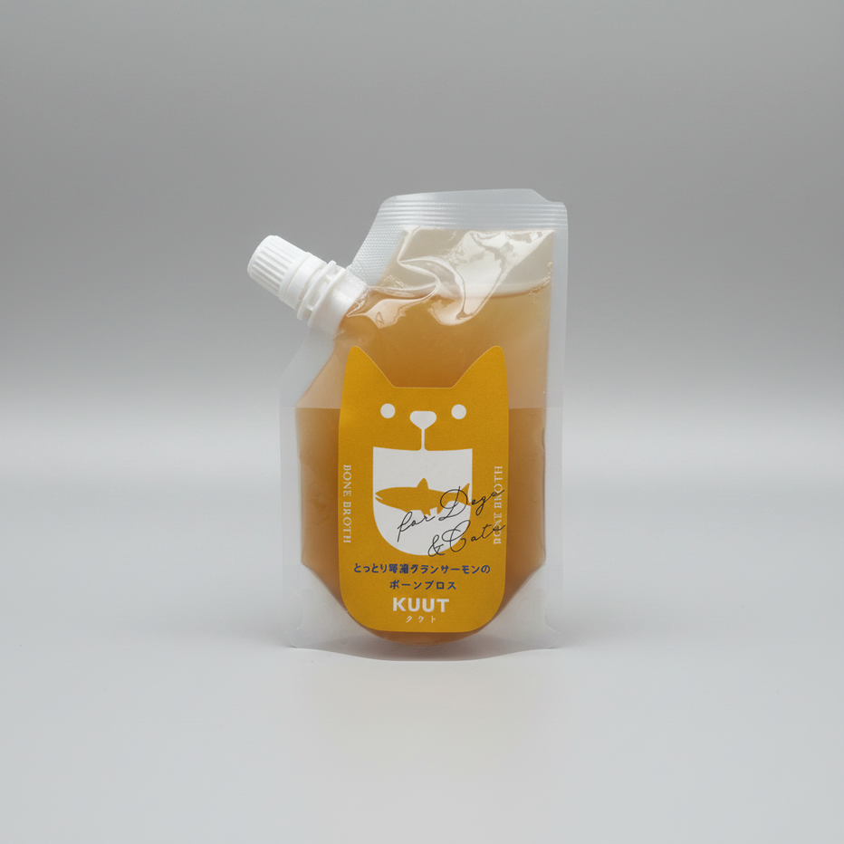 A squeeze pouch of KUUT Salmon flavour bone broth, filled with orange-coloured contents, featuring a yellow cat-shaped label with a fish illustration and white cap, set against a neutral grey background.
