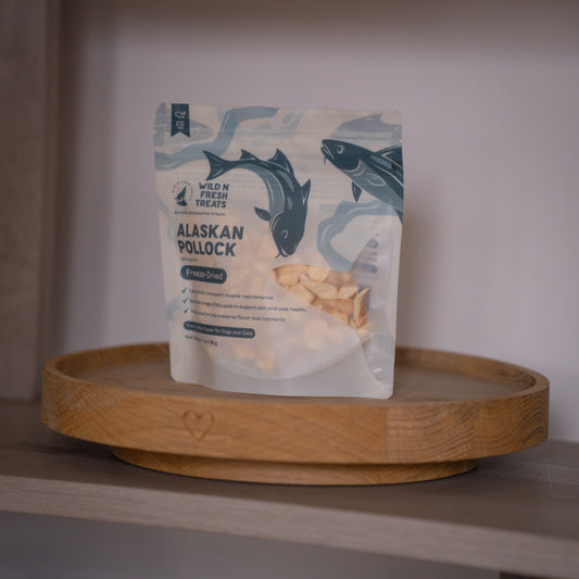 A stand-up pouch of Wild N Fresh Treats Alaskan Pollock freeze-dried treats, displayed on a round wooden tray against a neutral indoor background.
