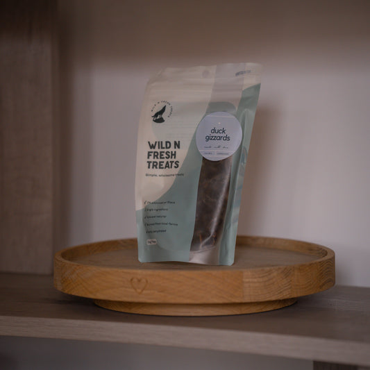 A stand-up pouch of Wild N Fresh Treats duck gizzards displayed on a round wooden tray against a neutral indoor background.