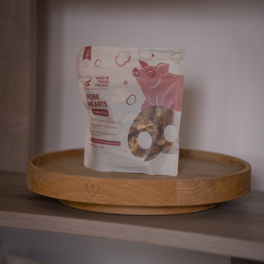 A stand-up pouch of Wild N Fresh Treats Pork Hearts dog treats sits on a round wooden tray against a soft neutral indoor background.