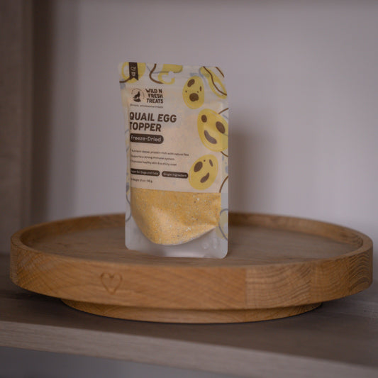 A stand-up pouch of Wild & Fresh Treats Quail Egg Topper freeze-dried pet food, standing on a round wooden tray against a neutral indoor background.